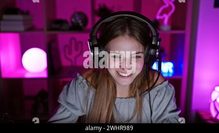 A smiling young woman with headphones enjoys gaming in a vibrant purple-lit room at night. Stock Photo