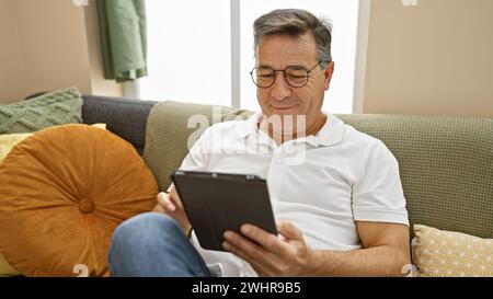 A middle-aged man relaxes at home, engaging with a tablet in his cozy living room. Stock Photo