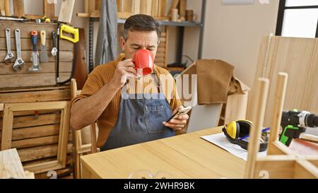 Mature man drinking coffee in a woodworking workshop while holding a smartphone, surrounded by tools and wooden materials. Stock Photo