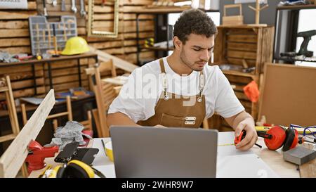 A young man with a beard measuring wood in a carpentry workshop, tools scattered around and laptop on the workbench. Stock Photo