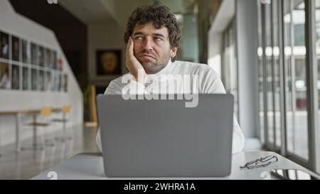 A pensive hispanic man with a beard sits at a desk in an office, looking away from his laptop. Stock Photo