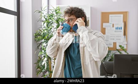 A young hispanic man sips coffee while on a phone call in an office, embodying casual professionalism. Stock Photo