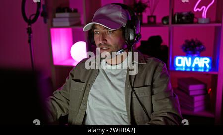 A young bearded man with headphones in a dark indoor gaming room, featuring neon lights and an 'on air' sign. Stock Photo