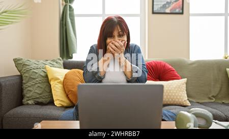 Shocked middle-aged woman covering mouth while using a laptop in a cozy living room. Stock Photo