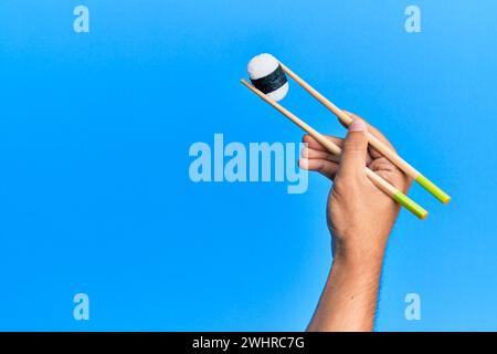 Hand of man holding sushi with chopsticks over isolated blue background Stock Photo