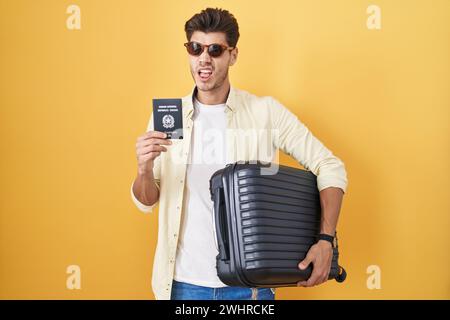 Young hispanic man holding suitcase going on summer vacation holding italian passport clueless and confused expression. doubt concept. Stock Photo