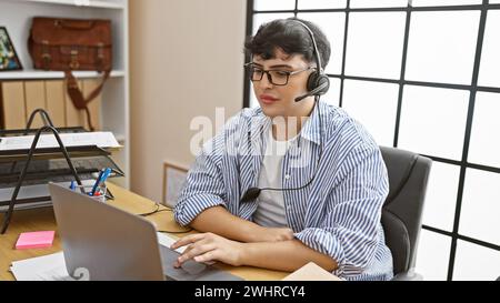 A young man in a striped shirt works diligently at his office desk, using a laptop and wearing a headset. Stock Photo
