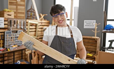 A smiling young man wearing safety glasses and an apron holds a piece of wood in a well-equipped carpentry workshop. Stock Photo