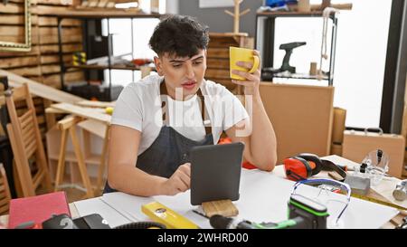 A young man examines his tablet while holding a cup in a well-equipped carpentry workshop. Stock Photo