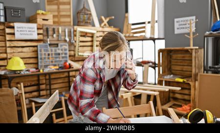 A focused young woman carpenter in casual attire is on a phone call while writing notes indoors at a woodworking workshop. Stock Photo
