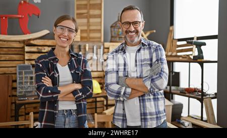Two confident carpenters, man and woman, beaming as they stand together with arms crossed in professional woodwork business, symbol of partnership and Stock Photo