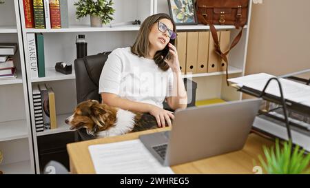 A professional young woman talks on the phone while petting her dog at her organized office space. Stock Photo