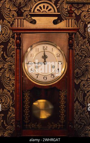 Vintage clock with pendulum on the wall. Nostalgia concept. Antique clock with ticker. Old fashioned clock face with arrows. Classic room design. Stock Photo