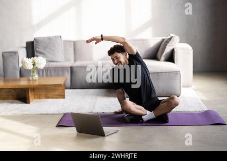 Active young cheerful man doing morning gymnastic at home, sitting on yoga mat and stretching, copy space Stock Photo
