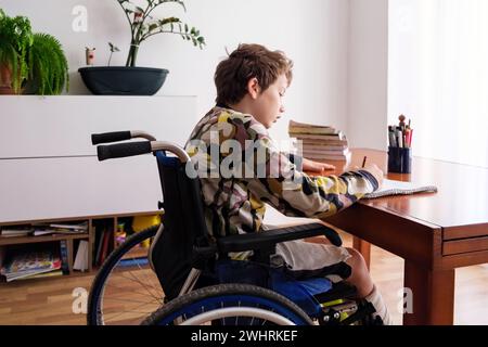 A boy in a wheelchair is seen sitting at a desk, focused on doing his homework. Stock Photo
