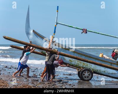 Traditional Indonesian fishing boat with outriggers being hauled onto a stony beach in Medewi on the island of Bali in Indonesia. Stock Photo