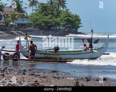 Traditional Indonesian fishing boat with outriggers being hauled onto a stony beach in Medewi on the island of Bali in Indonesia. Bombora Medewi Resor Stock Photo