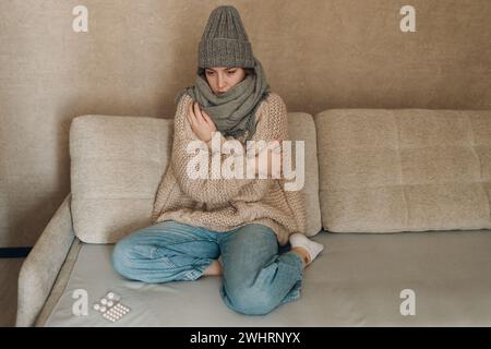 Young woman wearing scarf and wool hat has flu ill sick disease cold at home indoor on sofa. Stock Photo