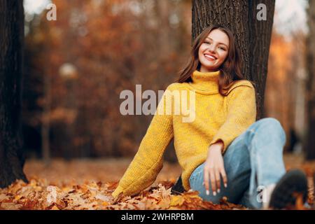 Beautiful girl at autumn park sitting near a tree with falling yellow foliage leaves. Woman smile Stock Photo