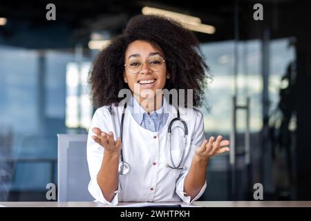Successful female surgeon in professional uniform raising hands while sitting by desk with toothy smile. Positive black woman ready announcing good news and positive prognosis of treatment results. Stock Photo