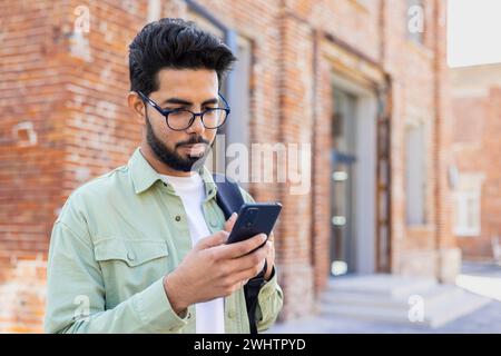 Young indian guy in eyeglasses looking at smartphone screen while standing with backpack outdoors. Focused young man checking online classes schedule for attending courses in university campus. Stock Photo
