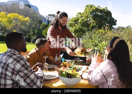 Happy diverse male and female friends serving thanksgiving celebration meal in sunny garden Stock Photo