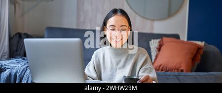 Close up portrait of beautiful asian woman drinks her coffee, smells drink in cup, takes brake from working or studying on lapto Stock Photo