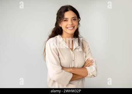 Smiling young woman standing confidently with arms crossed over light background Stock Photo
