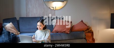 Vertical shot of asian girl sits on floor at home, working on laptop, studying at cozy place, using computer to freelance Stock Photo