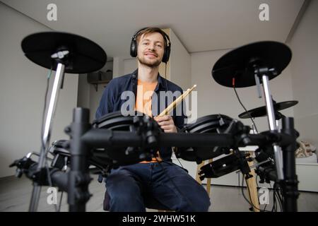 Portrait of smiling young man with headphones hitting electric drum set during repetition at home. Online music lessons. Stock Photo