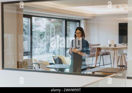A thoughtful professional woman enjoys a peaceful coffee break alone in a contemporary and well-lit office environment, reflecting on her workday. Stock Photo