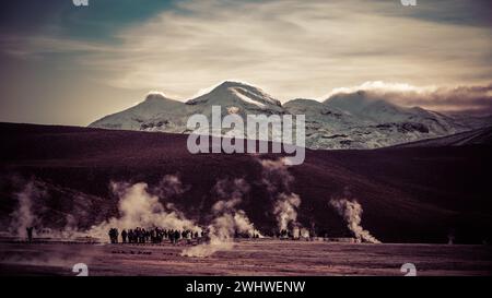 Snow-covered peak in the golden light of sunrise and a crowd of people amid clouds of steam from hot springs. Stock Photo