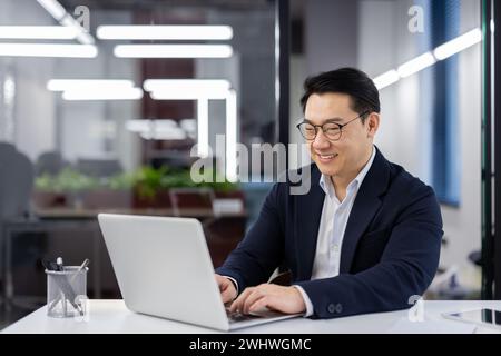 Asian man in business suit and glasses working with laptop inside office, mature businessman typing on keyboard, preparing financial report, male company employee at workplace. Stock Photo