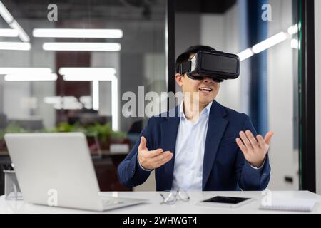 Businessman uses virtual reality glasses VR simulator, inside office at workplace, man in business suit, boss online meeting communicates with colleagues remotely and virtual world, smile joyfully Stock Photo