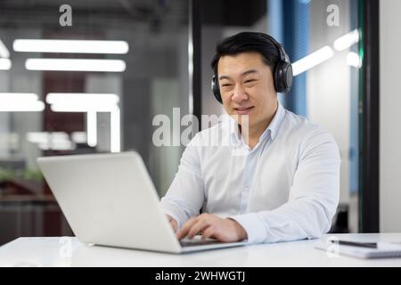Successful asian programmer with laptop inside office, mature experienced man in headphones listening to music, audio books and podcasts, worker in shirt using laptop to code new software. Stock Photo