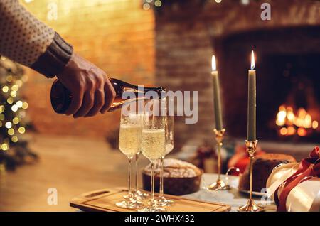 New Years Eve celebration. Man pouring champagne into glasses standing on table with festive xmas dinner, candles and wrapped gi Stock Photo