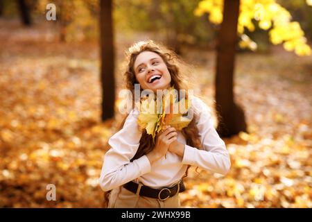 Smiling young woman enjoying the autumn weather in the forest with the yellow leaves in the hands at sunset. Stock Photo