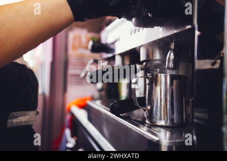 Barista in black gloves presses the button for pouring boiling water into metal cup while preparing coffee drink or cocktail. Stock Photo
