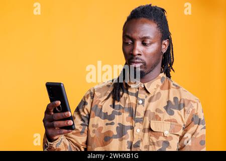 Young adult typing message using mobile phone, posing over yellow background in studio. African american man holding smartphone browsing on internet, reading new online article Stock Photo