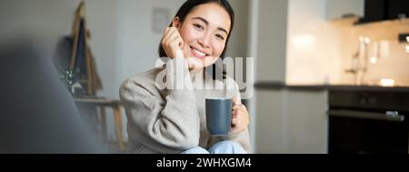 Smiling asian woman sitting at home with cup of coffee, relaxing and feeling warmth, looking outside window, resting on sofa in Stock Photo