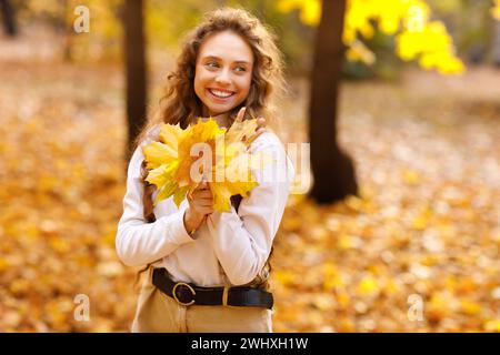 Smiling young girl enjoying the autumn weather in the forest with the yellow leaves in the hands at sunset. Stock Photo