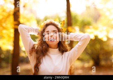 Smiling young woman with the fall crown of yellow leaves on her curly hairstyle head in the autumn park at sunset. Stock Photo