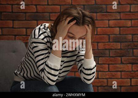 Sad young woman sitting on chair near brick wall, space for text Stock Photo