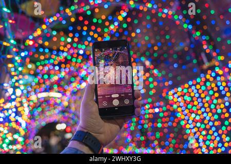 Male hand holding a smartphone and taking a picture with colorful bokeh Christmas lights in the background Stock Photo