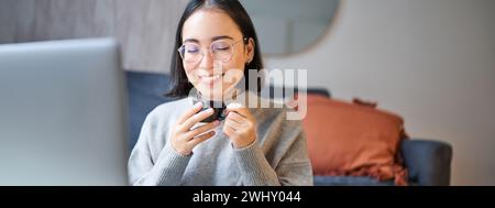 Close up portrait of beautiful asian woman drinks her coffee, smells drink in cup, takes brake from working or studying on lapto Stock Photo