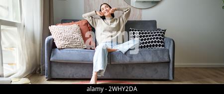 Portrait of happy asian woman feeling lazy, stretching on sofa and smiling pleased, relaxing at home, resting from work Stock Photo