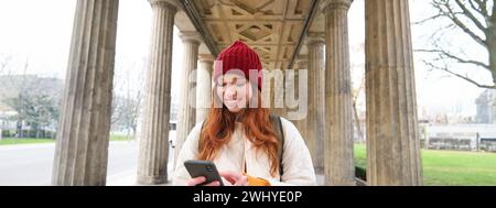 Mobile broadband and people. Smiling redhead 20s girl with backpack, uses smartphone on street, holds mobile phone and looks at Stock Photo