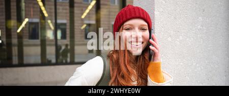 Young people and mobile connection. Happy redhead girl talks on phone, makes telephone call, stands outdoors with backpack and u Stock Photo