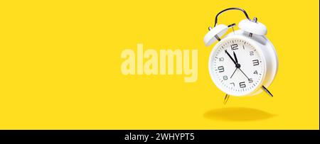 White bell alarm clock hovering over yellow background. 5 to 12 clock concept isolated. Stock Photo