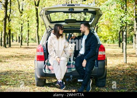 A young couple enjoying a serene moment, sipping beverages sitting in the trunk of their car, accompanied by a white fluffy dog Stock Photo